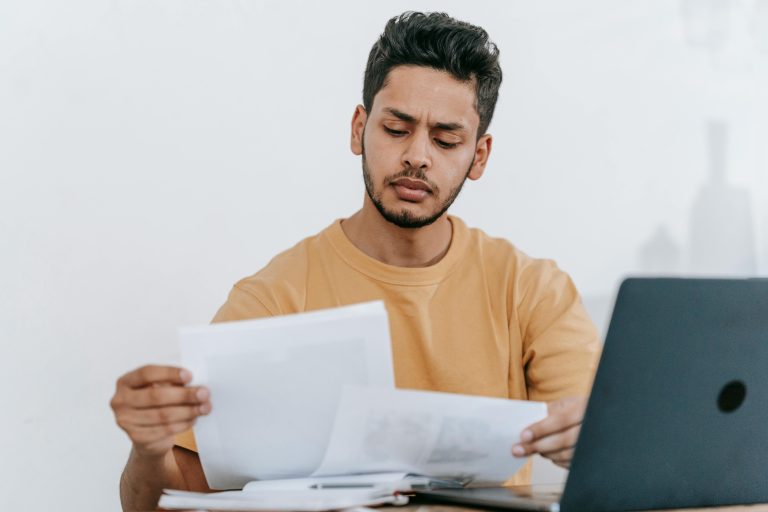 Focused young man reviewing paperwork at his desk, showcasing a business setting with a laptop indoors.